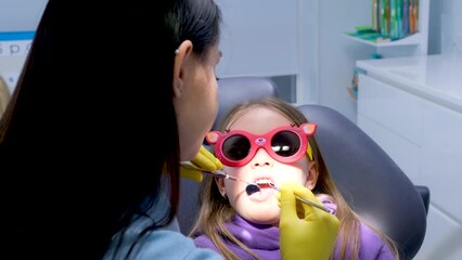 pediatric dentistry close-up little girl opened mouth shows teeth doctor looks at whether she has caries fillings treatment examination by doctor girl wears red sunglasses so that dental lamp