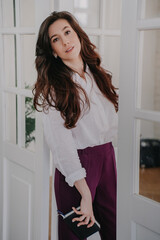 Calm brunette woman with wavy dark hair in white shirt and violet pants standing in living room, looking at camera, holds diary. Tired businesswoman home after busy day at work. Business people.
