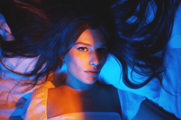 Fototapeta premium Portrait, vaporwave lights and young woman face in bedroom with creative disco lighting. Makeup, beauty and model resting and feeling relax and calm on a bed pillow with cyberpunk aesthetic