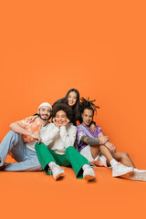 multiethnic friends in trendy clothes sitting and smiling at camera on orange background.