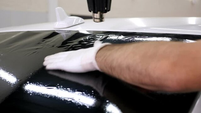 The master in gloves installs a tinting film on the car glass with a hair dryer and a spatula in bright light. Conceptual tinting of the car.