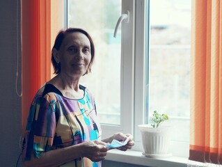 A portrait of an older woman with deep wrinkles in quarantine at home, holding a medical mask. Grandmother stands and looks out the window