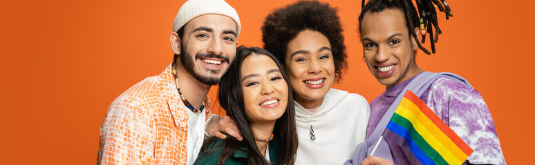 trendy multiracial friends smiling at camera near lgbt flag isolated on orange, banner.