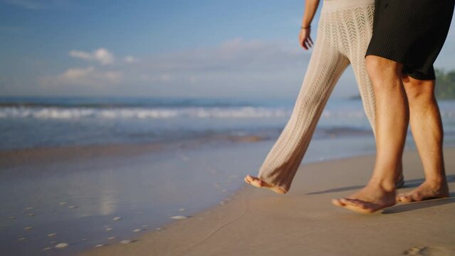 Legs of happy couple holding hands and walking on the beach together enjoying summer side view. Feet of heerful boyfriend and girlfriend relaxing and taking a walk at the seaside at sunrise side shot.