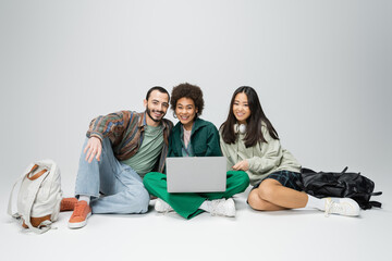 full length of happy and trendy multicultural students sitting with laptop on grey background.