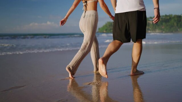 Legs of happy couple holding hands and walking on the beach together enjoying summer back view. Feet of heerful boyfriend and girlfriend relaxing and taking a walk at the seaside at sunrise back shot.