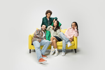 happy multiracial friends in trendy outfit posing on yellow couch on grey background.