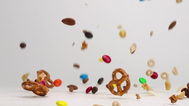 Sweet and salty trail snack mix with peanuts, pecan, almond, and cashew nuts, pretzels, m and m candies and cheerios falling onto white table top in slow mo