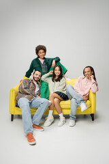 fashionable multiethnic friends posing on yellow couch and smiling at camera on grey background.
