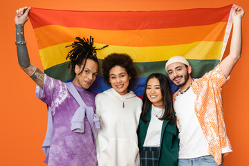 cheerful and trendy multiethnic friends posing with lgbt flag isolated on orange.