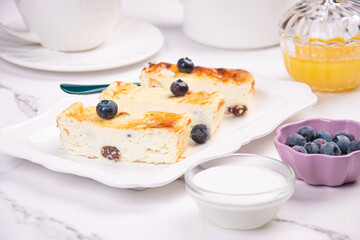 Cottage cheese casserole on a white plate on a white table. Served with blueberries, sour cream and honey.