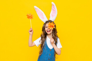 Happy Easter holiday. A beautiful little girl with rabbit ears on her head, celebrating a spring day. A child in a sundress holds origami flowers on a yellow isolated background.