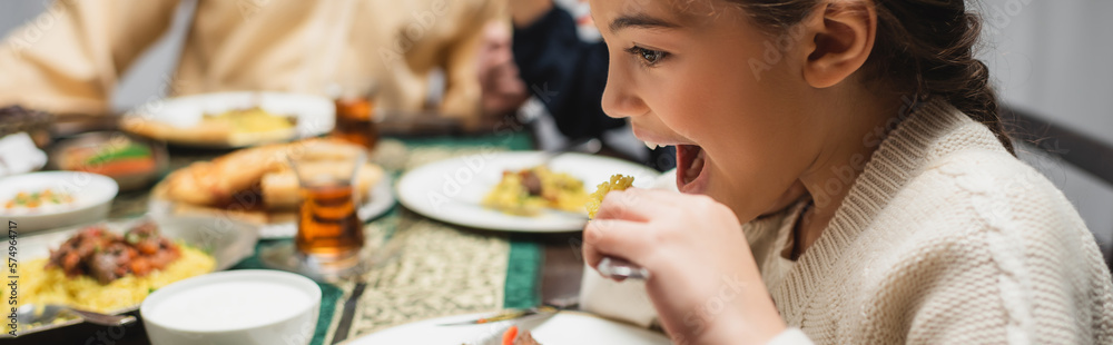 Wall mural Muslim girl eating iftar dinner near blurred food and parents at home, banner.