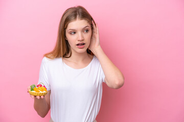 Teenager Russian girl holding a tartlet isolated on pink background listening to something by putting hand on the ear