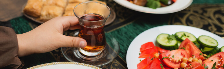 Cropped view of muslim boy holding traditional glass cup of tea near iftar food at home, banner.