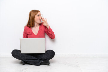 Young redhead woman with a laptop sitting on the floor isolated on white background shouting with mouth wide open to the lateral