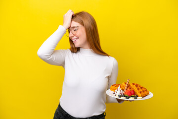 Young redhead woman holding waffles isolated on yellow background has realized something and intending the solution