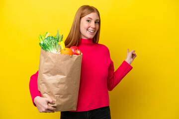 Young redhead woman holding a grocery shopping bag isolated on yellow background pointing back