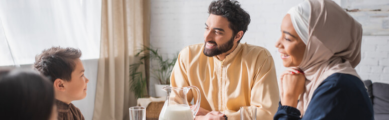 Fototapeta premium Positive middle east parents looking at kids near milk at home, banner.