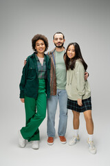 full length of young bearded man embracing happy multiethnic women on grey background.