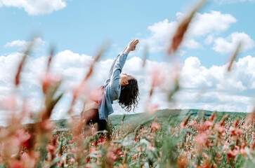 Woman doing standing yoga backbend with arms up in flower field with bright cloudy sky