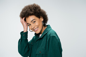 pleased african american woman in stylish shirt touching curly hair and looking at camera isolated on grey.