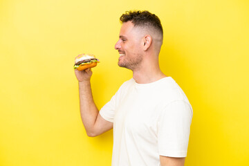 Young caucasian man holding a burger isolated on yellow background laughing in lateral position