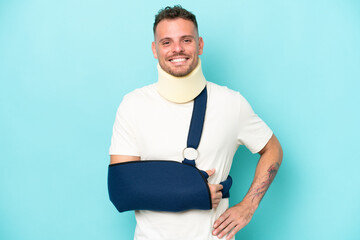 Young caucasian man wearing a sling  and neck lace isolated on blue background posing with arms at...