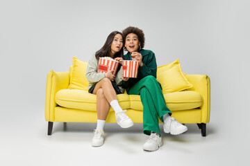 full length of astonished interracial women holding popcorn while watching movie on yellow couch on grey background.