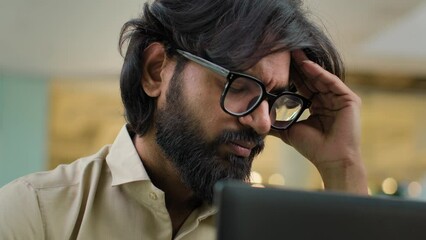 Close up worried stressed Indian bearded man in glasses suffer from head high pressure headache painful head thinking problem solution with laptop at workplace. Exhausted tired Arabian businessman