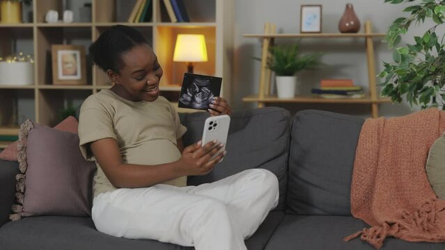 Smiling Pregnant African American Woman hatting with friends via Smartphone, Showing Ultrasound Scan of Baby During Video Call on Smartphone. Future Mother Enjoying Sonogram in Living Room