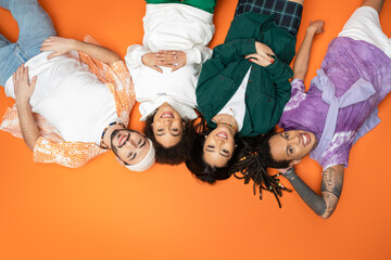 top view of happy multicultural friends in stylish outfit looking at camera while lying on orange background.