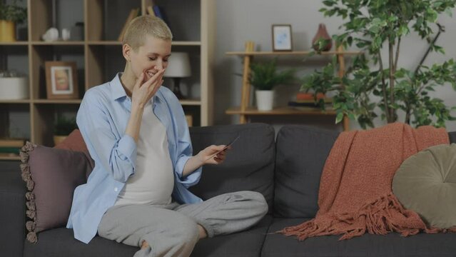 Attractive Pregnant Woman Sitting on Cozy Sofa Looking at Ultrasound Scan or Sonogram of Baby and Smiling. Female Blonde with Short Haircut Feeling Sincere Happiness During Expectation
