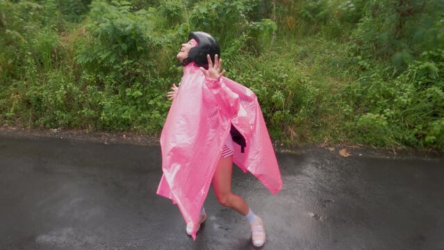 Cute Korean girl wearing bright neon pink raincoat dancing under rain on a wet road in rural area. Beautiful young woman with hiking bags and plastic Macintosh smiling while goofing and fooling.