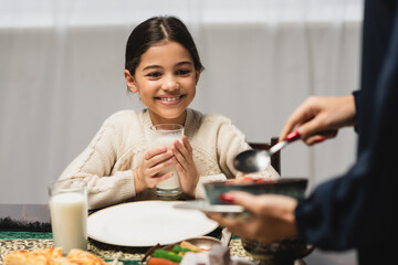 Smiling muslim girl holding glass of milk near blurred mom and ramadan dinner.