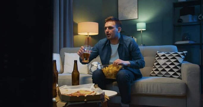 Man sitting on sofa at night in dark living room, eating potato chips and drinking beer. Male football fan is angry as favorite team loosing or missing goal. Man watching TV