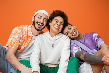 cheerful multiracial man looking at camera near friends laughing with closed eyes isolated on orange.