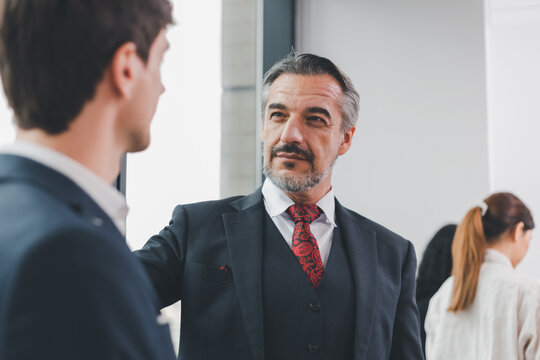 Two Caucasian businessmen wearing a formal suit, looking at each other eyes with pleasure smiling. The senior manager encourage a successful subordinate by touching his shoulders and shake the hand