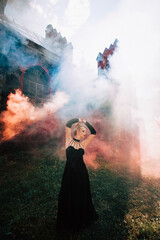Obraz premium Art work. A slender beautiful woman with blond hair in a black vintage dress and expensive jewelry, stylish makeup and hairstyle, comes out of an ancient castle in fire and smoke.
