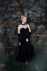 A slender beautiful woman with blond hair in a black vintage dress and expensive jewelry, stylish makeup and hairstyle, stands near the walls of an old castle.