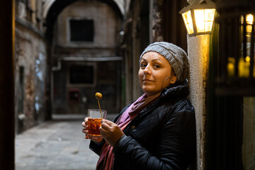 Fototapeta premium A woman enjoys a classic Italian aperitivo, savoring a Spritz drink in a cozy Venetian alley on a chilly evening, Venice, Italy