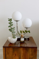 White vase with eucalyptus on a wooden pedestal. In the background there is a lamp