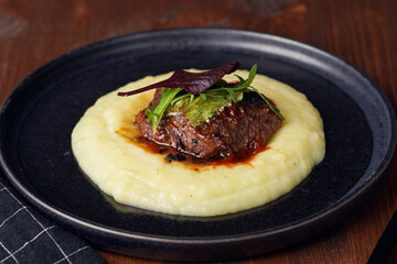 A medium rare ostrich steak in barbecue sauce on a cloud of herb mashed potato topped with fresh green arugula leaves on a black plate on a wooden background