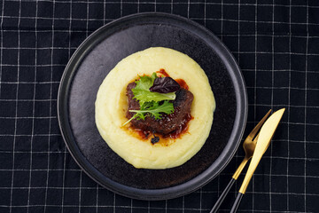 A medium rare ostrich steak in barbecue sauce on a cloud of herb mashed potato topped with fresh green arugula leaves on a black plate on a black checkered table cloth, top view