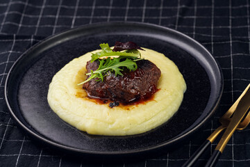 A medium rare ostrich steak in barbecue sauce on a cloud of herb mashed potato topped with fresh green arugula leaves on a black plate on a black checkered table cloth