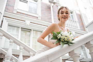 low angle view of happy bride in wedding dress holding bouquet and looking at camera near house.