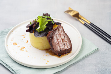 A medium rare ostrich steak in barbecue sauce with herb mashed potatoes topped with fresh green arugula leaves on a white plate on a grey background