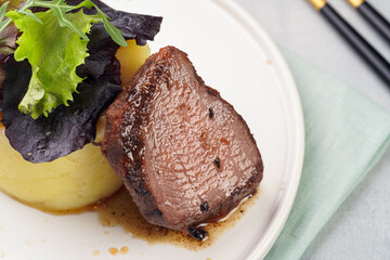 A medium rare ostrich steak in barbecue sauce with herb mashed potatoes topped with fresh green arugula leaves on a white plate on a grey background