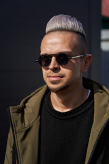 Obraz premium a young caucasian man with a white hair and sunglasses stands in a sunny color in a sweater and a green jacket and looks seriously