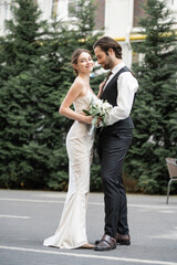 full length of bearded groom in vest embracing cheerful bride in white dress with wedding bouquet.